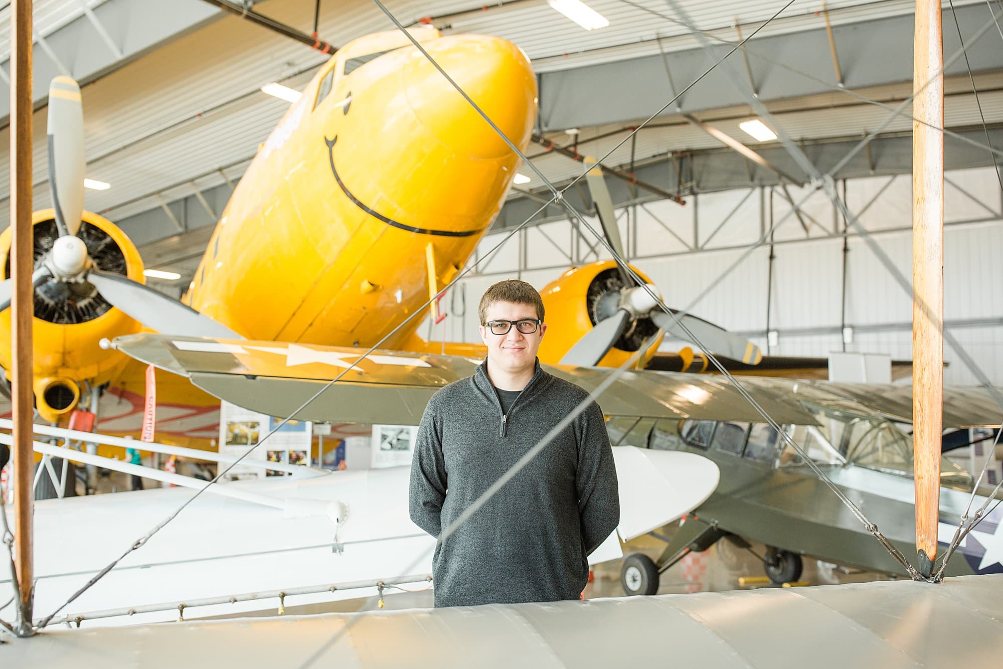 High school senior in grey sweatshirt smiles in front of a yellow plane during his Senior Session at Fargo Air Museum