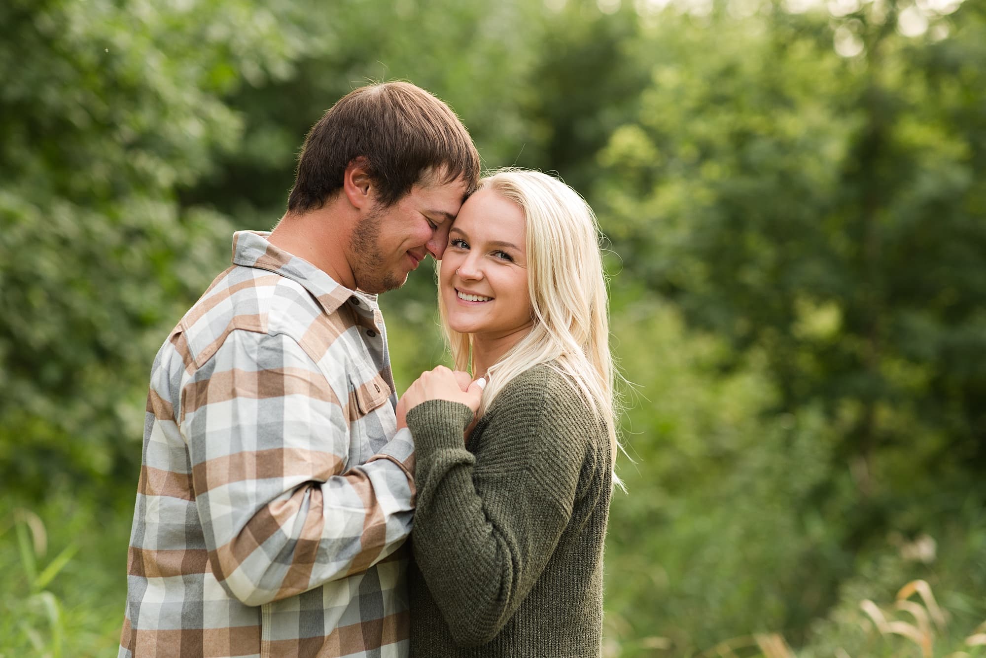 A woman in a green sweater smiles and holds hands with her fiance during their Engagement Session in Lindenwood Park