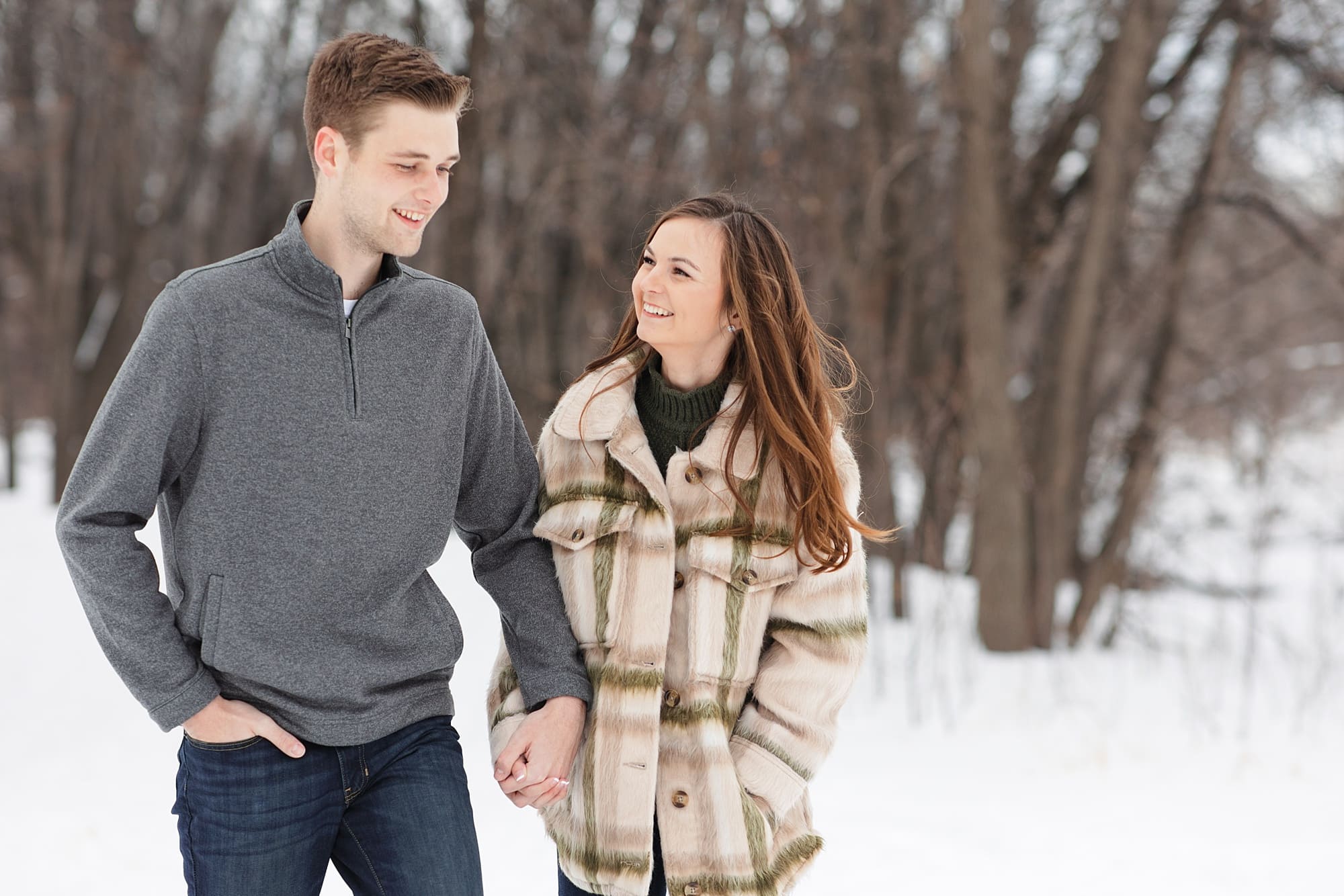 Engaged couple in grey sweater and tan jacket walk through the snow