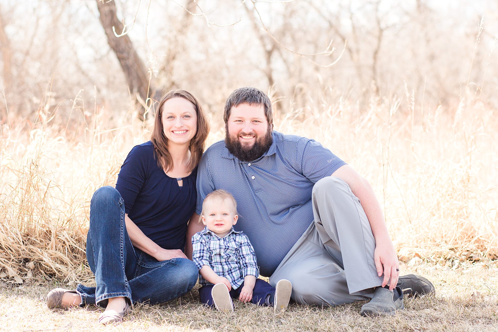 Family of three in shades of blue sit in tall grass with little boy celebrating his First Birthday