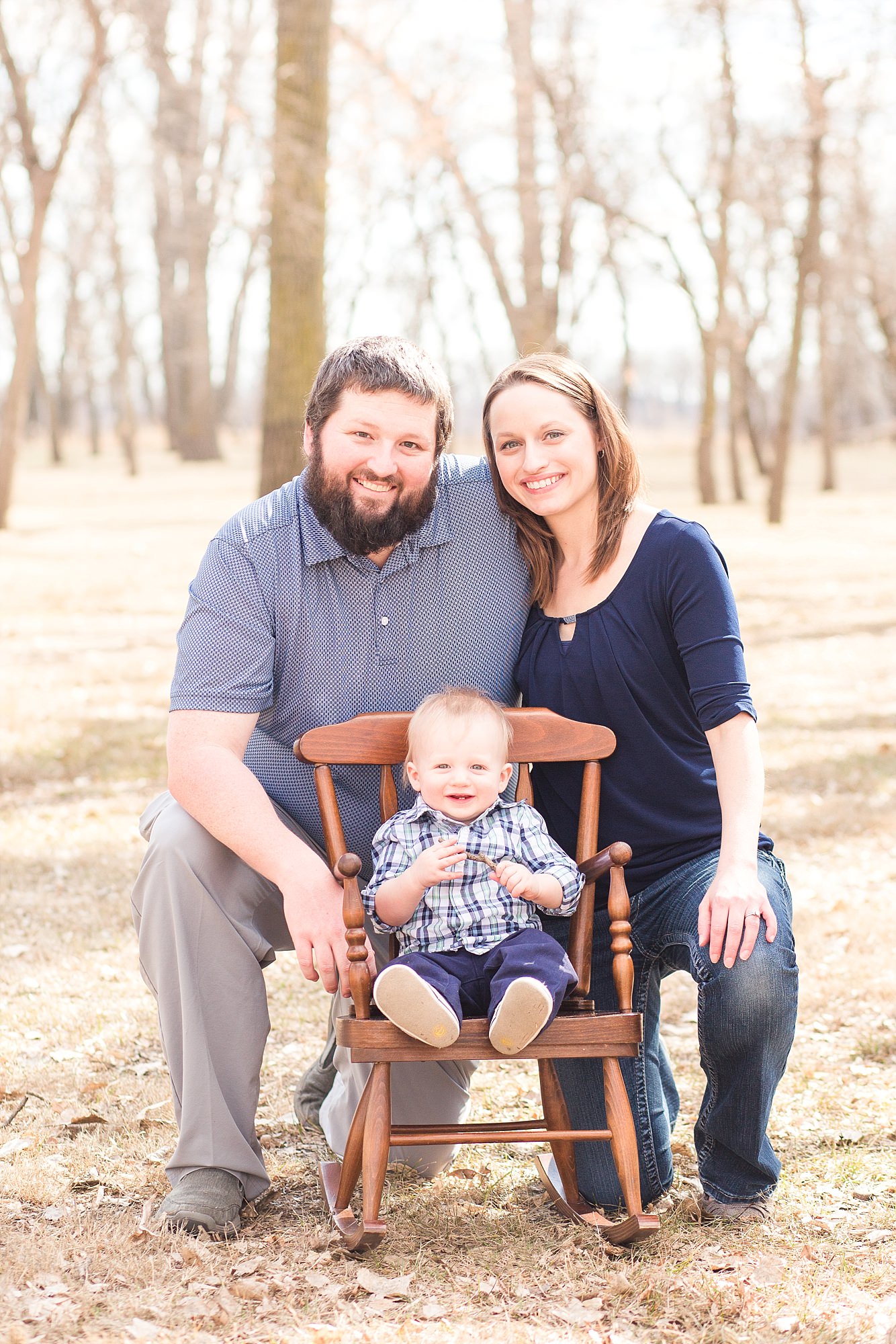 A family of three smile around a rocking chair with their son on his First Birthday