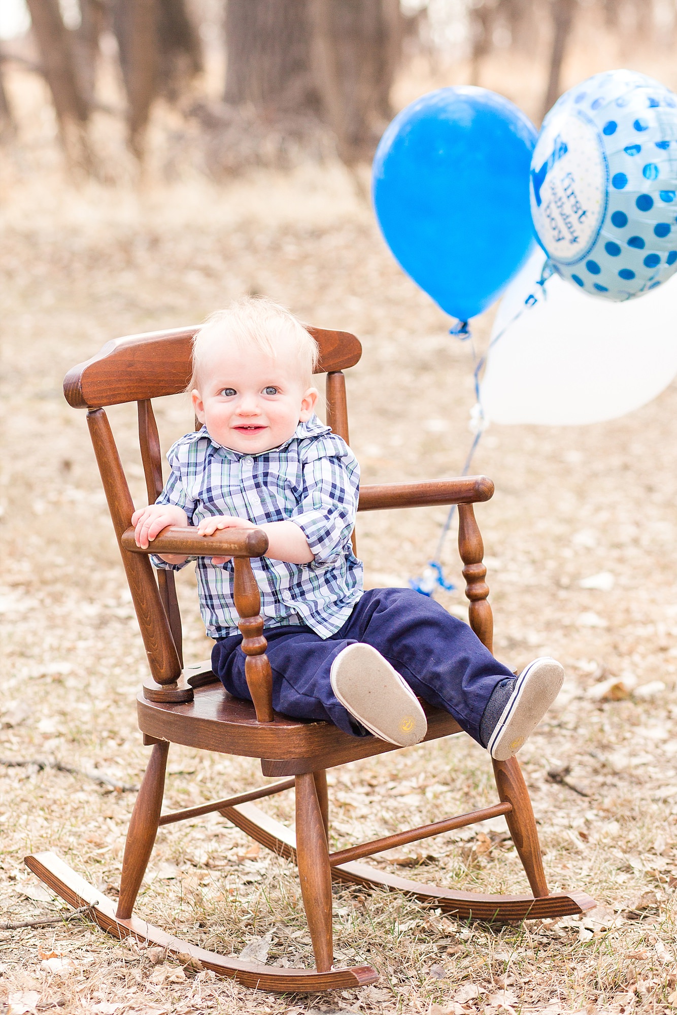 A First Birthday boy sits in a rocking chair with blue balloons blowing in the wind behind him