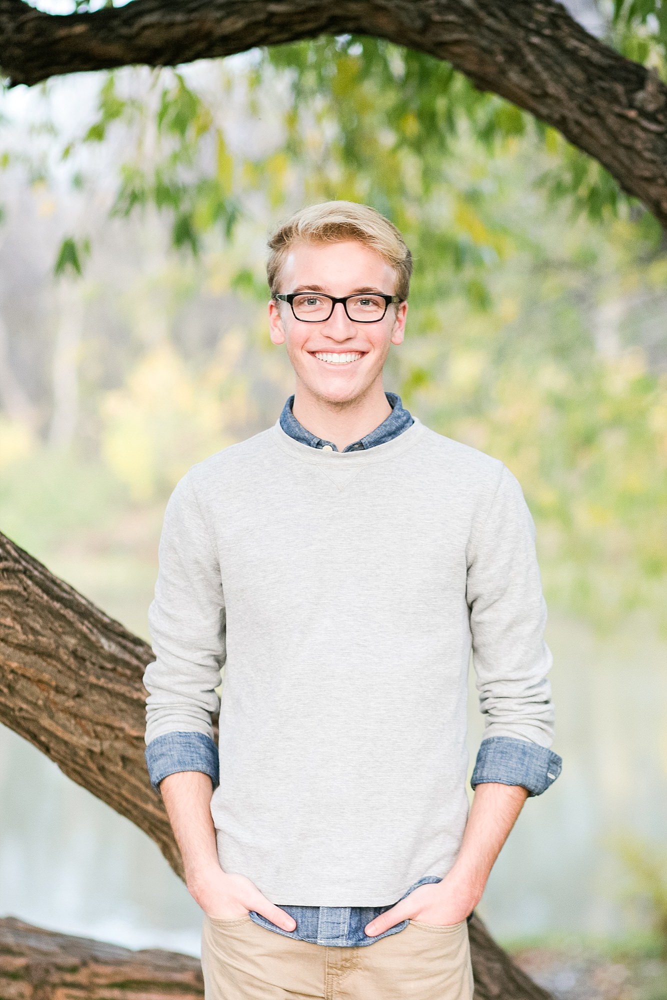 Senior guy smiles in front of the Red River, a tree, and green and yellow leaves in Fargo, ND