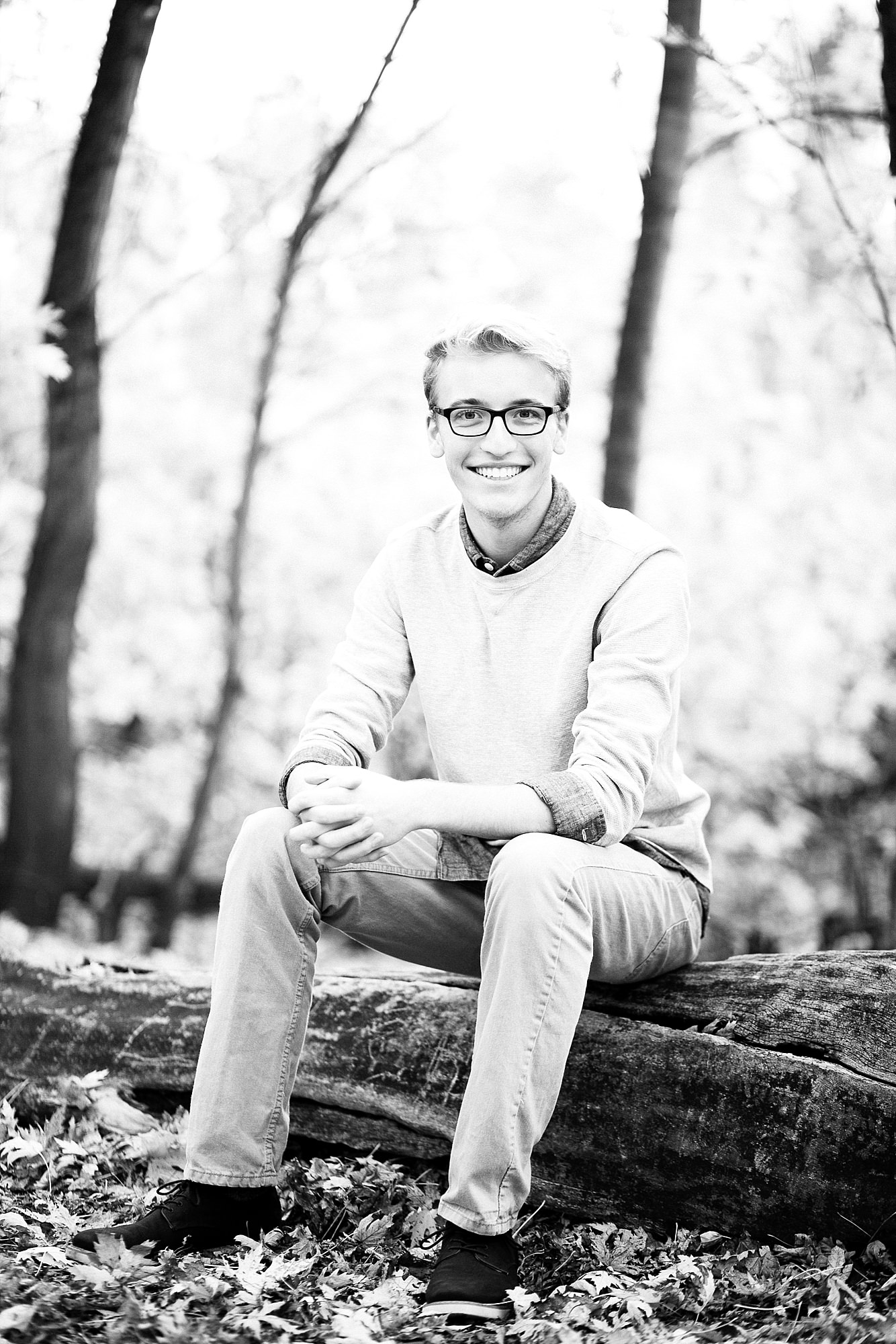 Senior in black and white sits on a tree in his grey sweater and black glasses