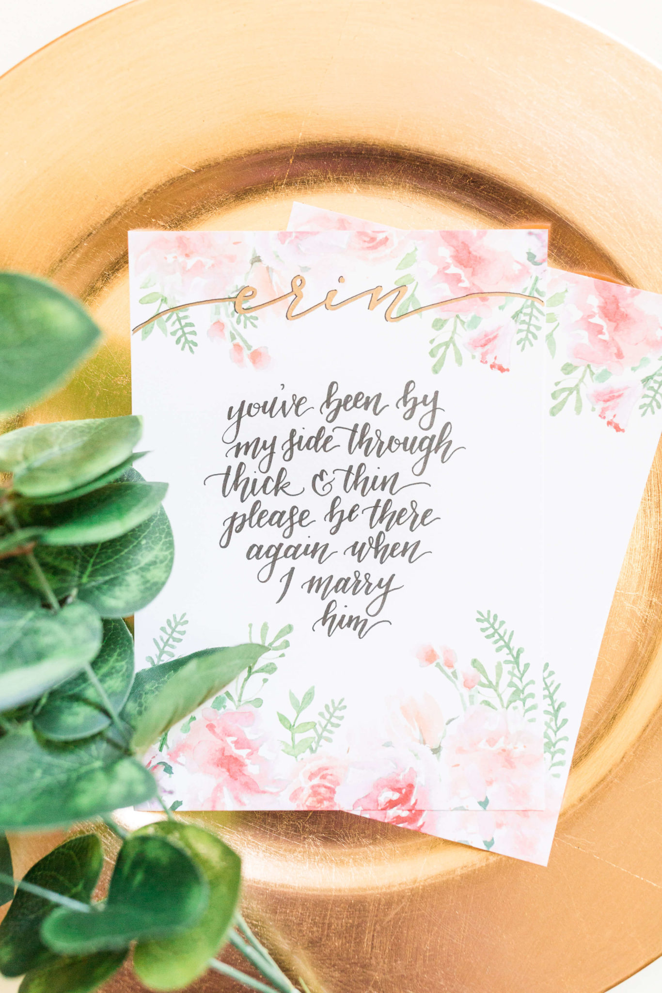 A floral bridal party invite lays on a gold plate with greenery