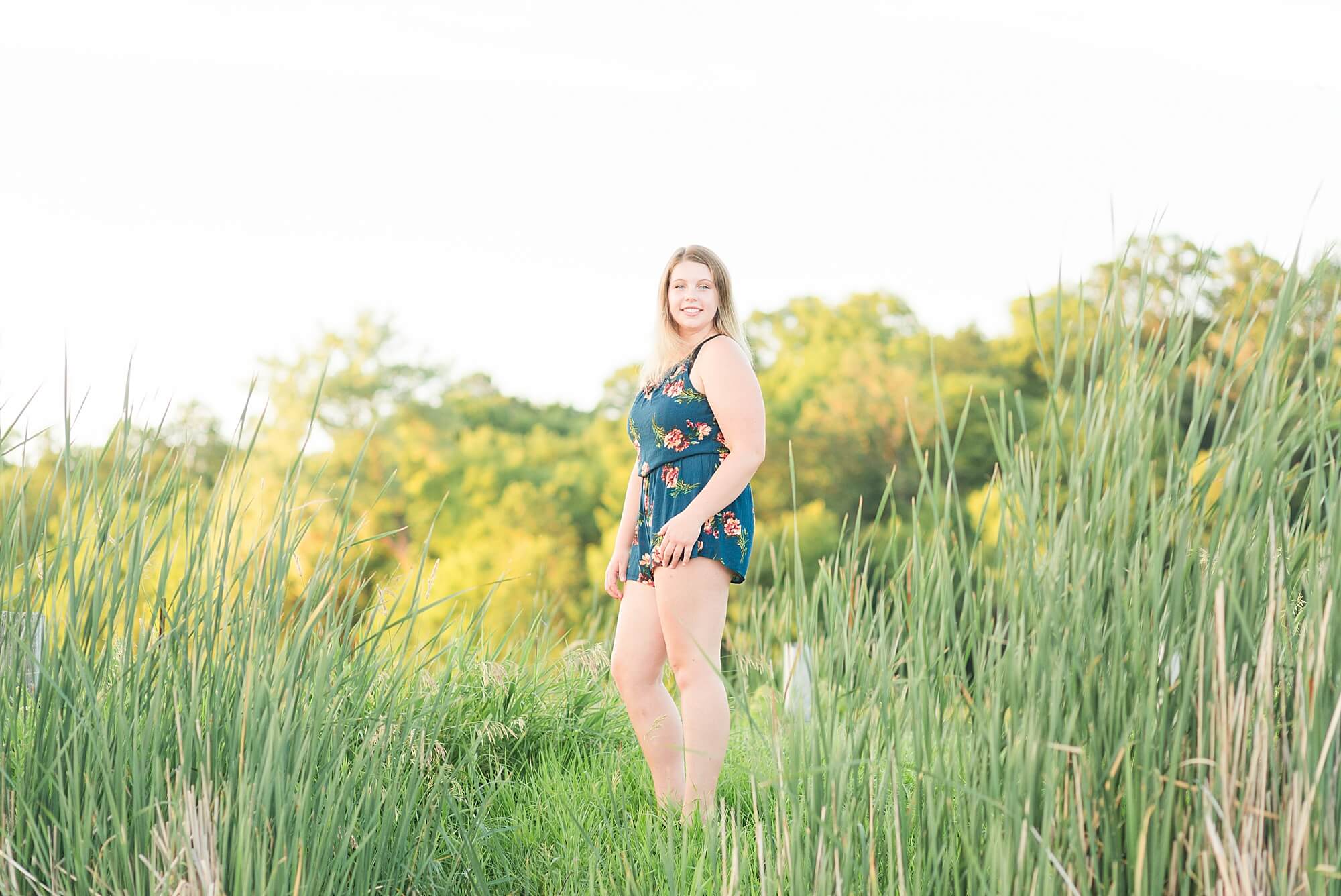 A teenager in a blue floral romper smiles along the lake and tall grass