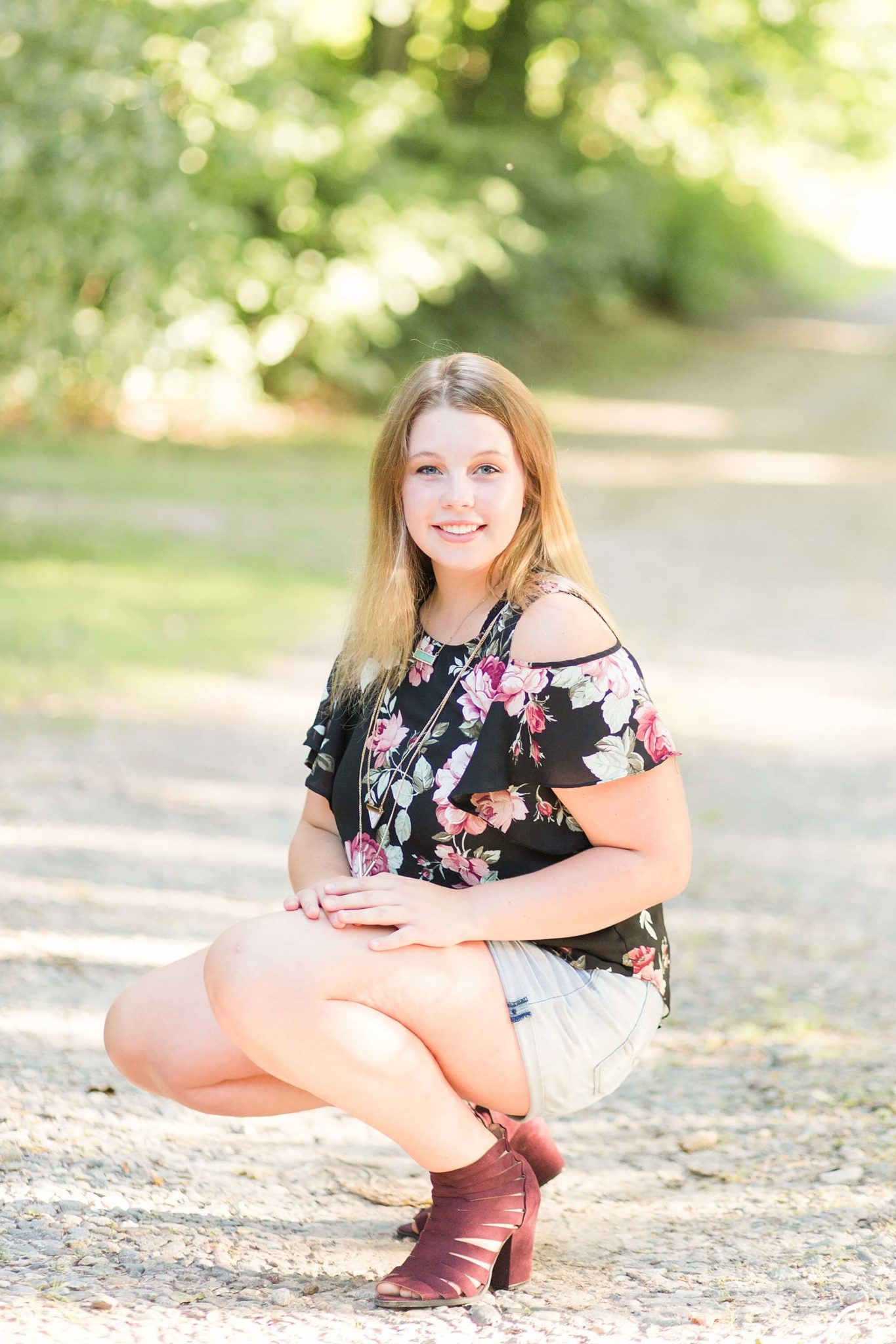 During a Lakeside Senior Portrait Session, a girl crouches down in maroon heels and a floral shirt.