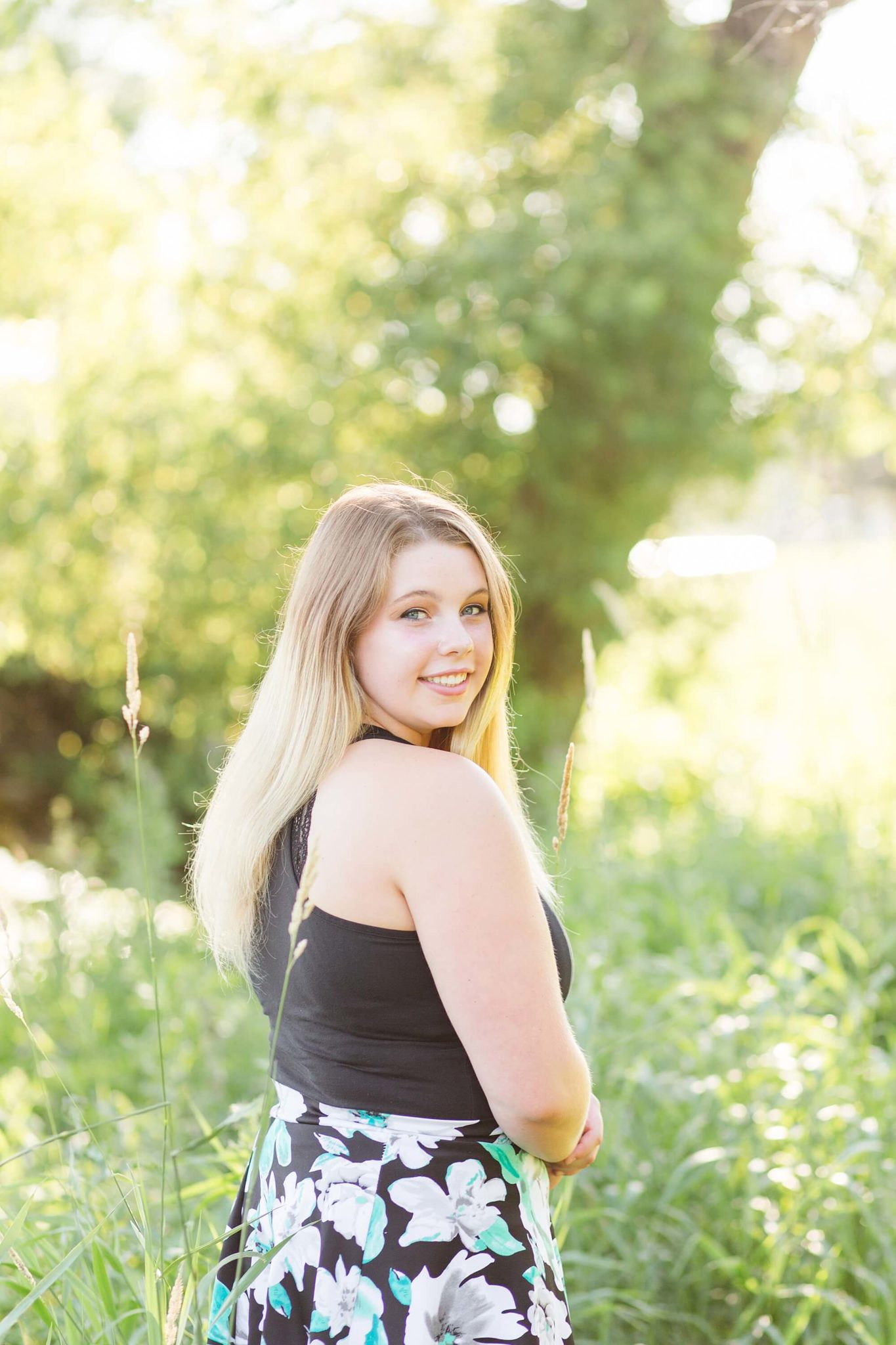 A senior girl smiles over her shoulder in the tall grass along the lake 