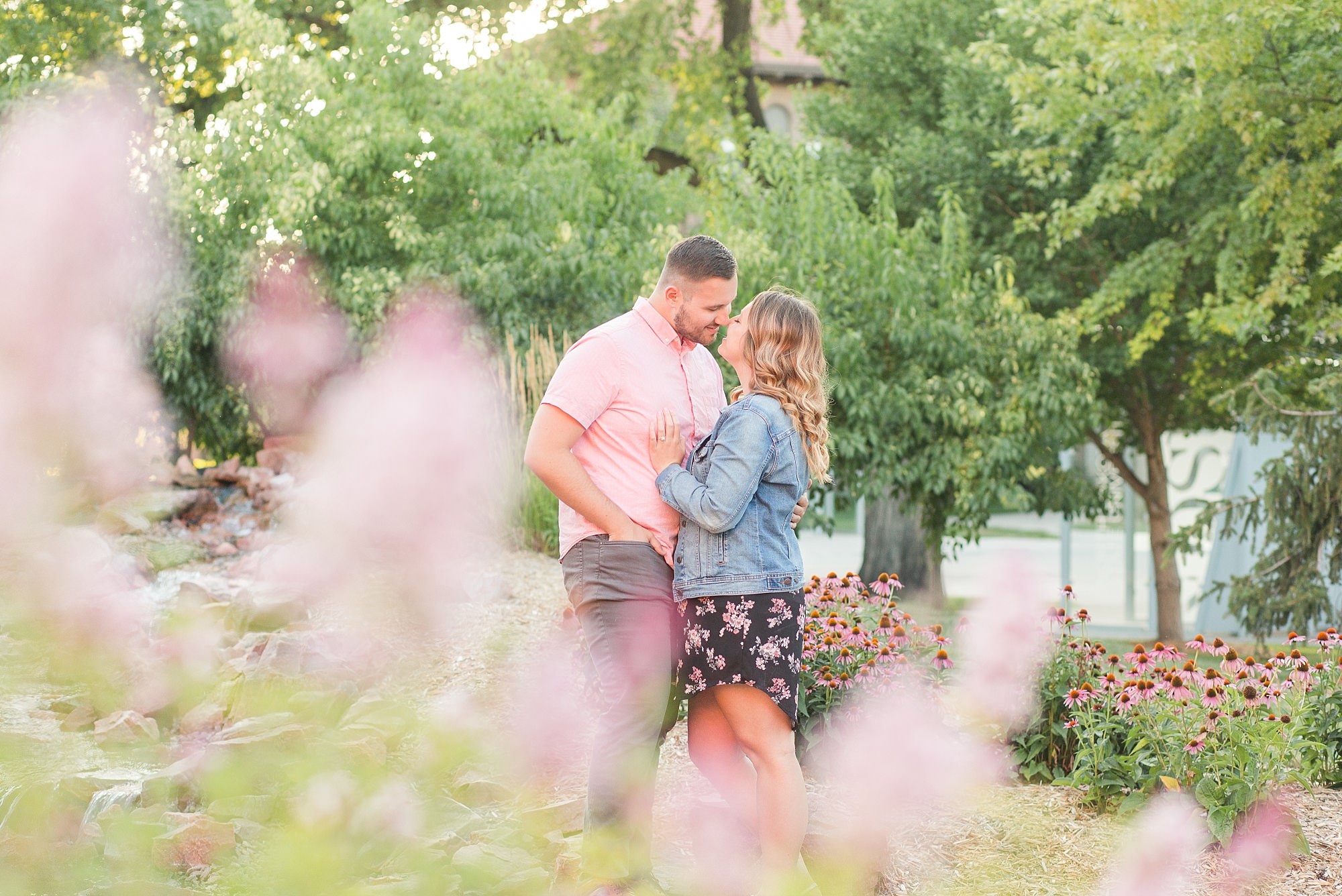 Engaged couple in pink, denim, and floral print kiss among pink and purple flowers