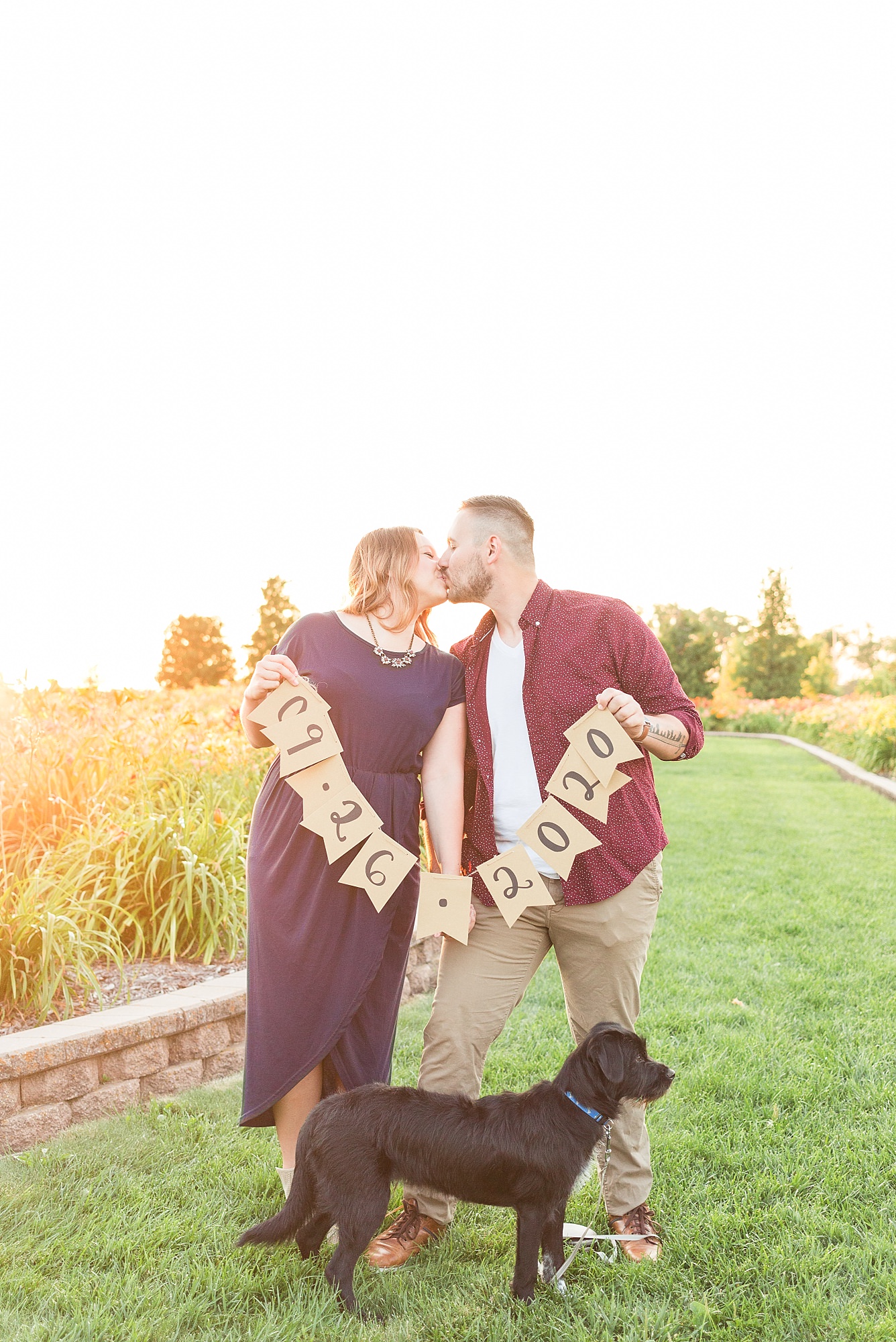 A couple kisses and holds a banner with their engagement date printed