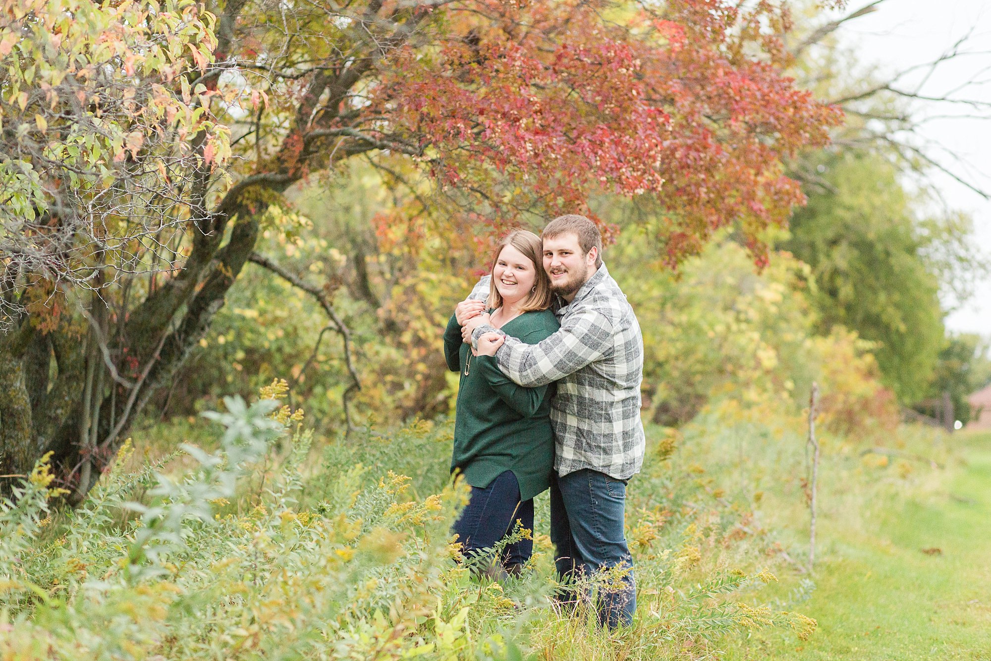 In green and grey plaid, an engaged couple smile in front of a red fall tree