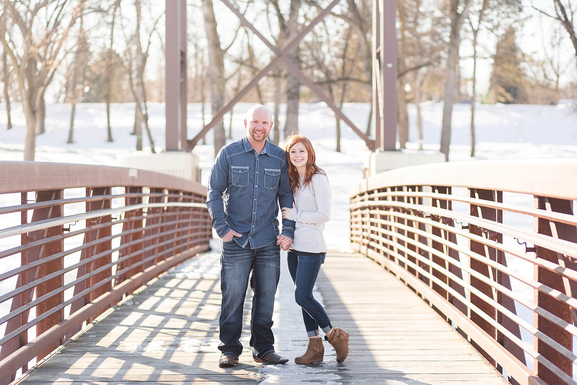 A tall man in blue and his redhead fiancee hold hands and smile on the Lindenwood bridge in Fargo