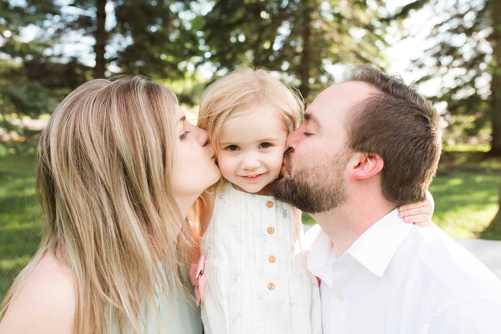 Young parents kiss their toddler on her checks during their adorable summer family session in Fargo, ND