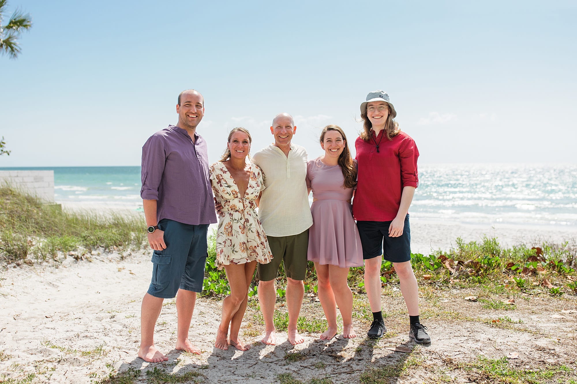 Adult Family of five smiling on the sandy beach with the blue ocean and sky behind them