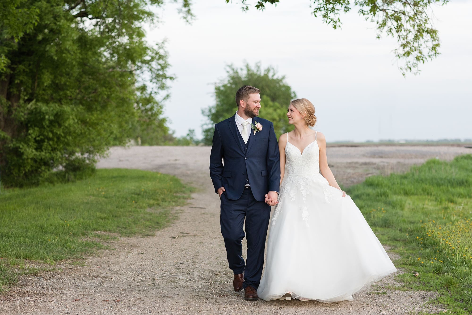 Bride and groom hold hands and walk along the gravel road at  Romantic Moons Event Center