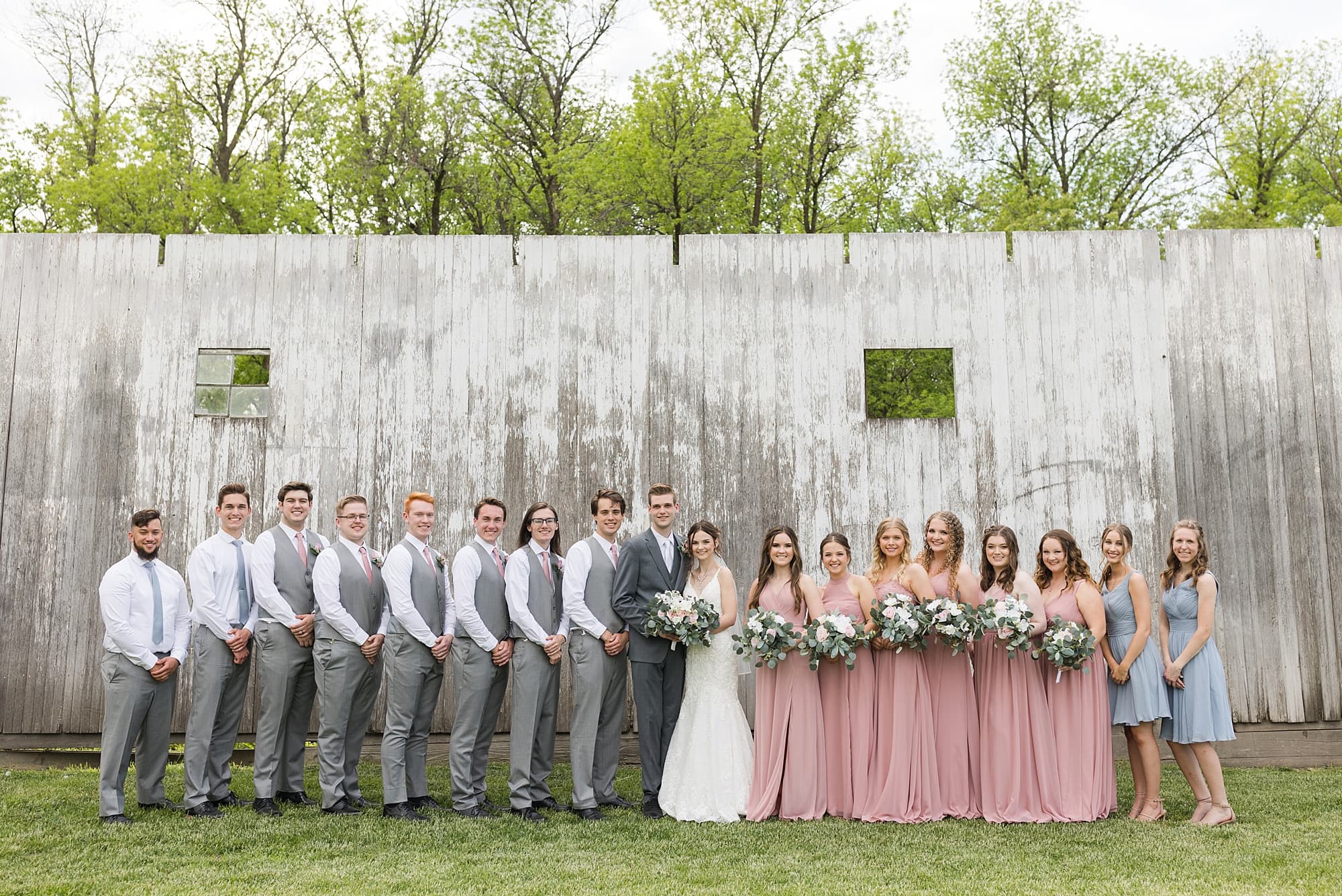 A large wedding party in dusty rose and grey smile at Legacy Acres Events
