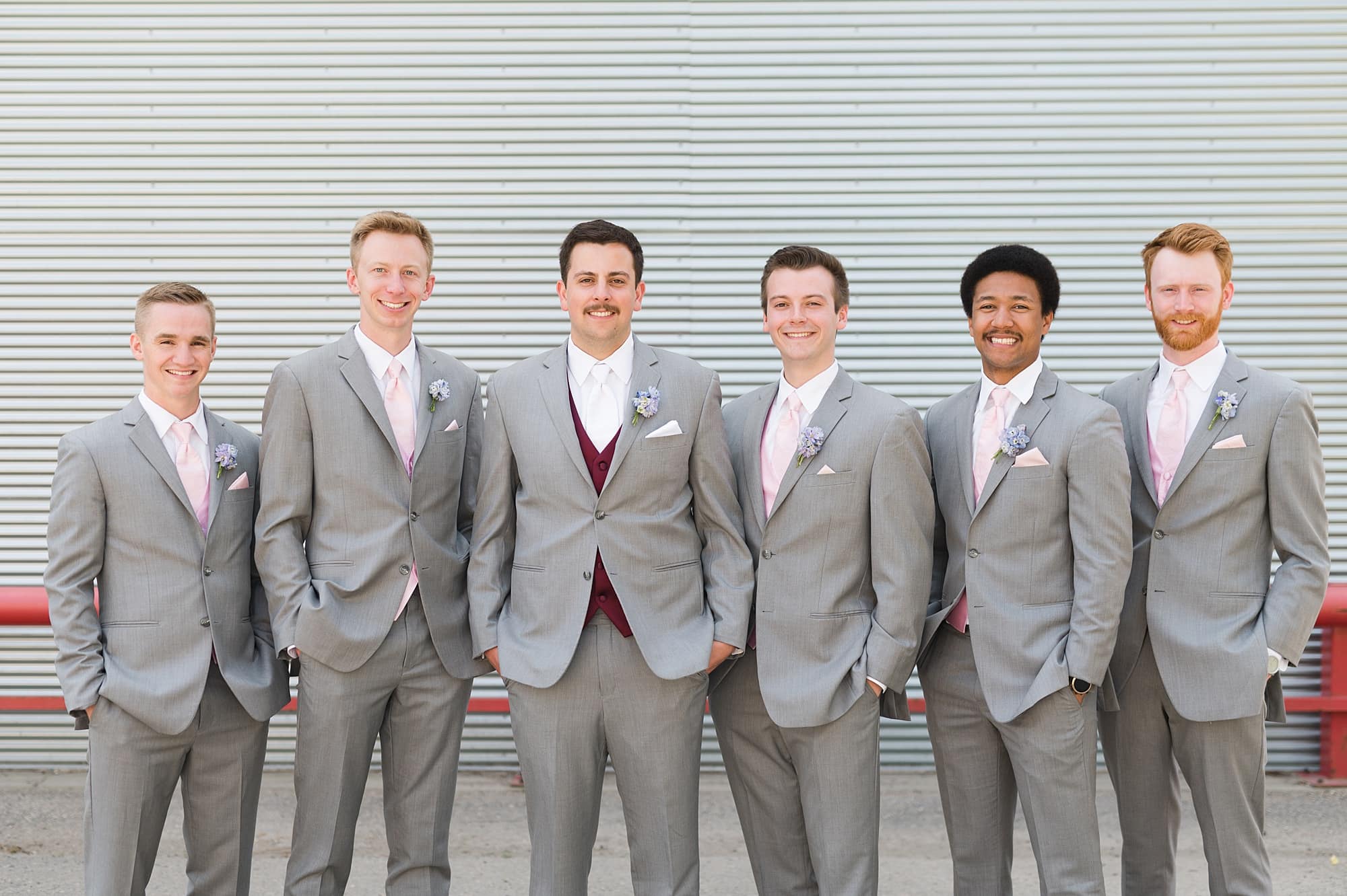 Groom and groomsmen in grey suits smile in front of a grey metal wall in Downtown Fargo
