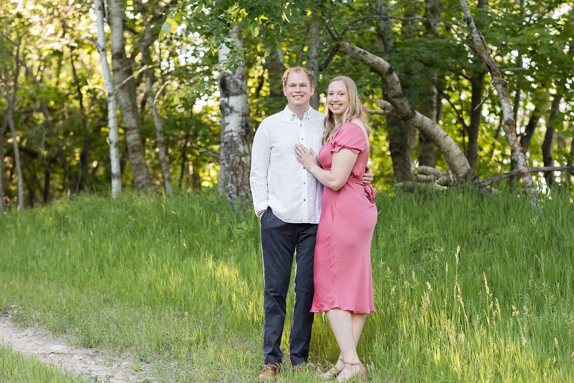 Engaged couple in light pink and white smile among the trees at Cotton Lake