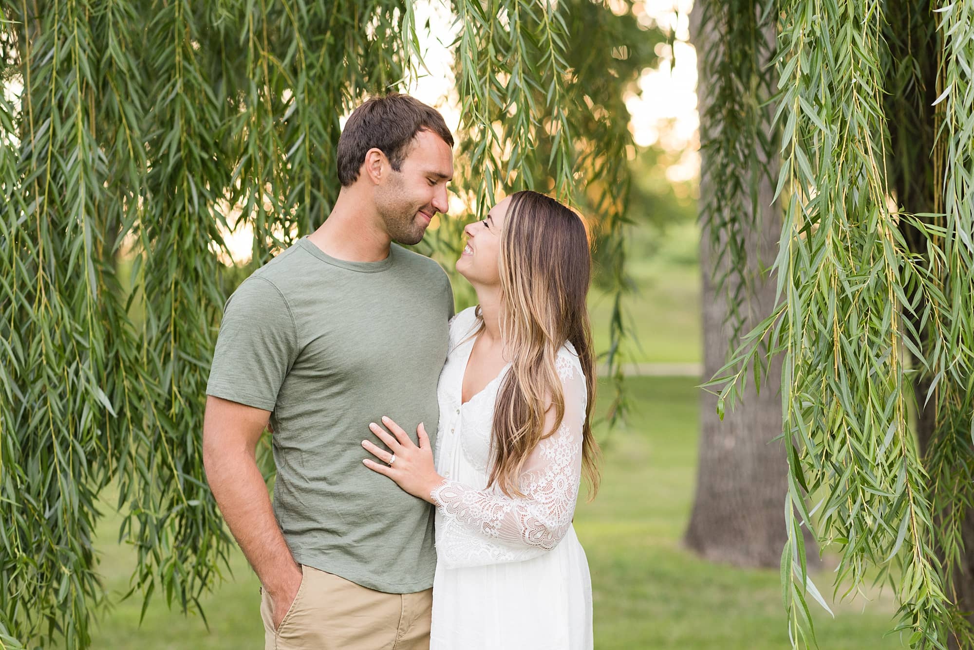 A couple smiles under a willow tree during their sunset engagement session in Lindenwood Park