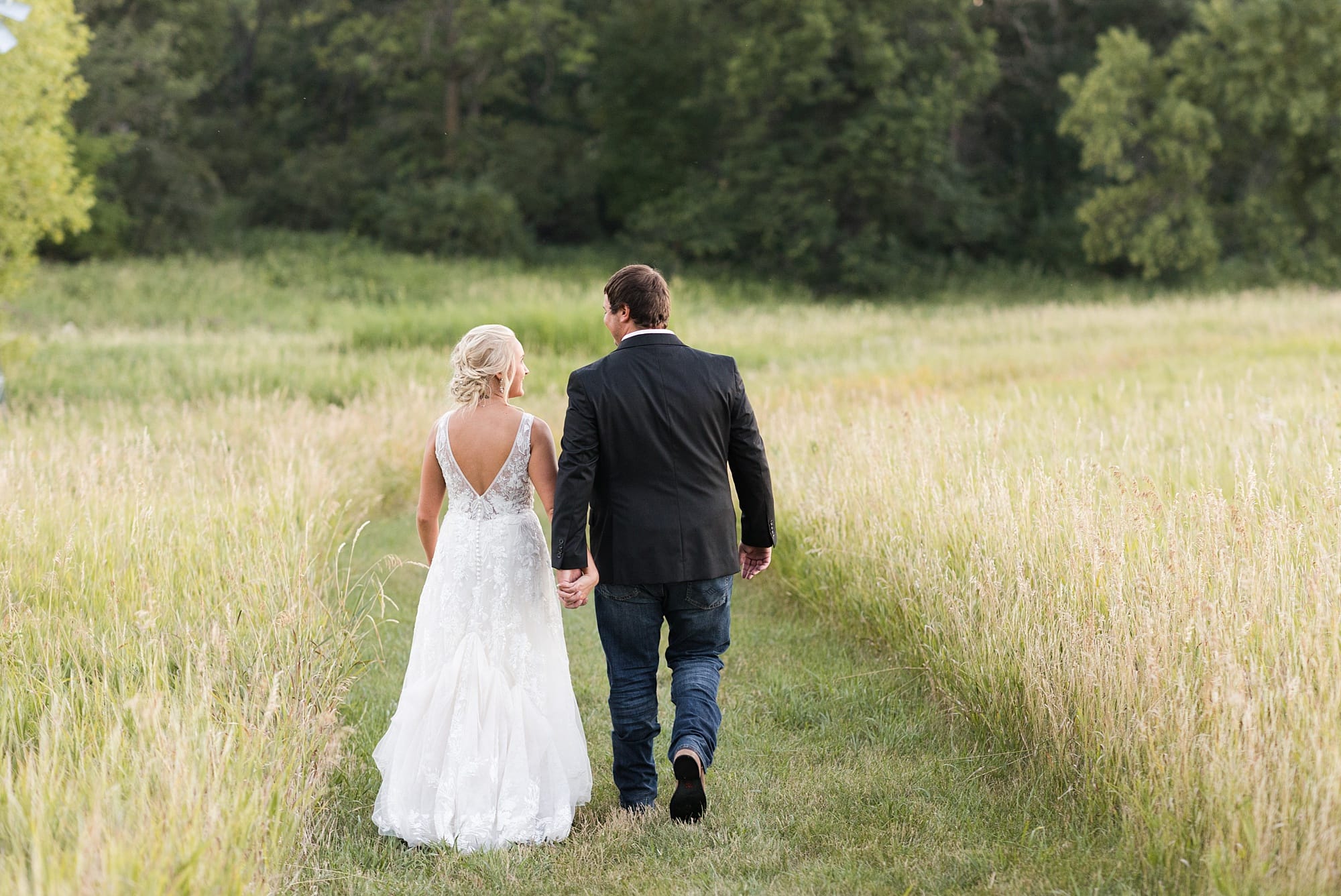 Bride and groom hold hands and walk down a grassy path during their  summer wedding at Rustic Oaks