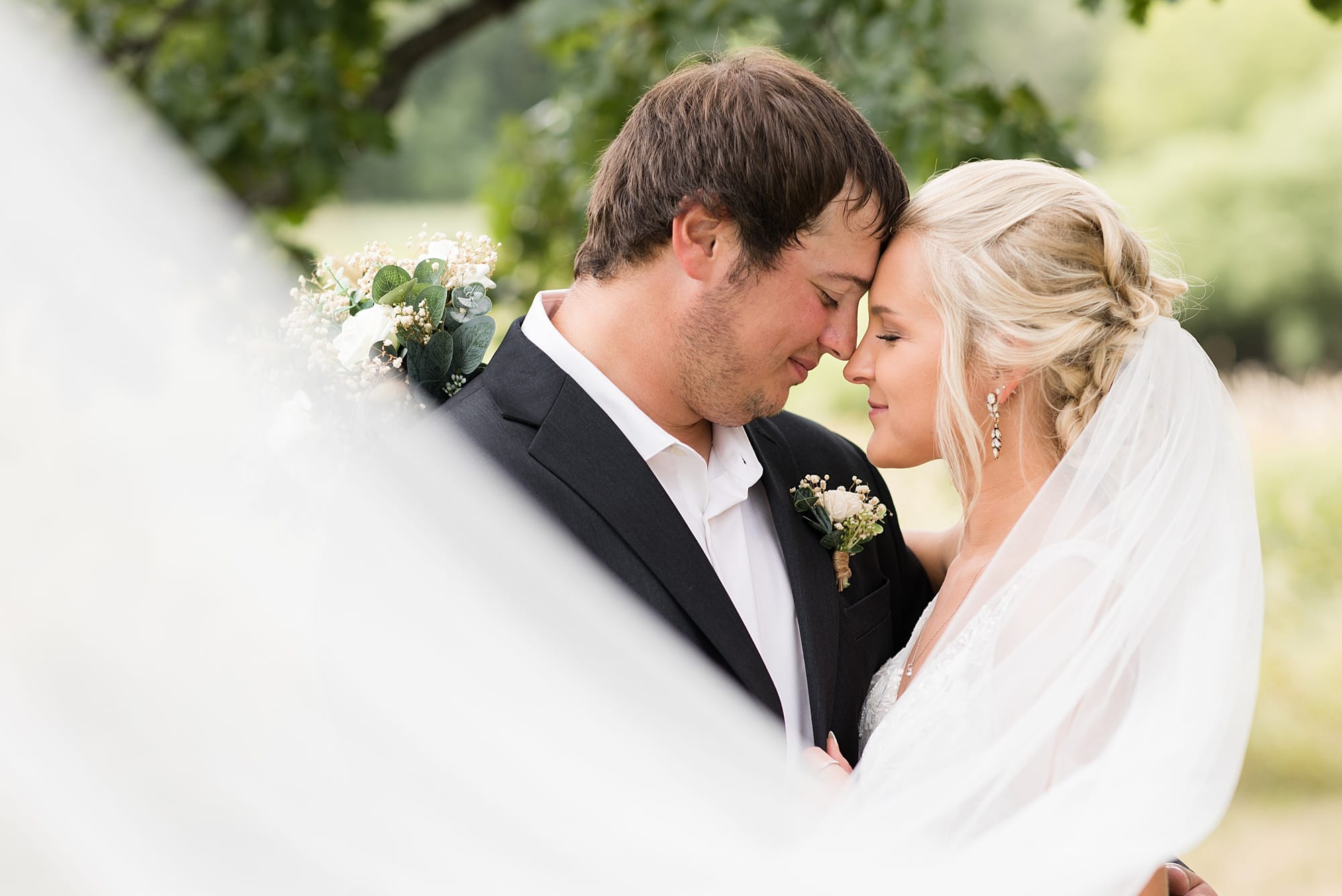 A bride and groom with a veil swoop close their eyes during their portrait session at Rustic Oaks