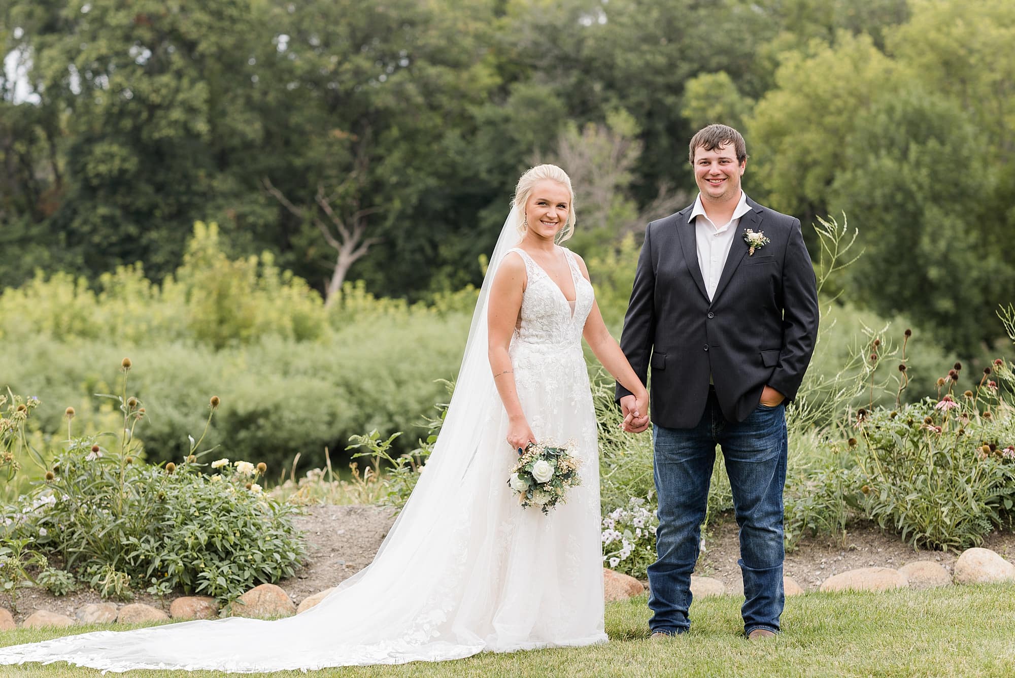 Bride and Groom smile and hold hands along the flowers at Rustic Oaks