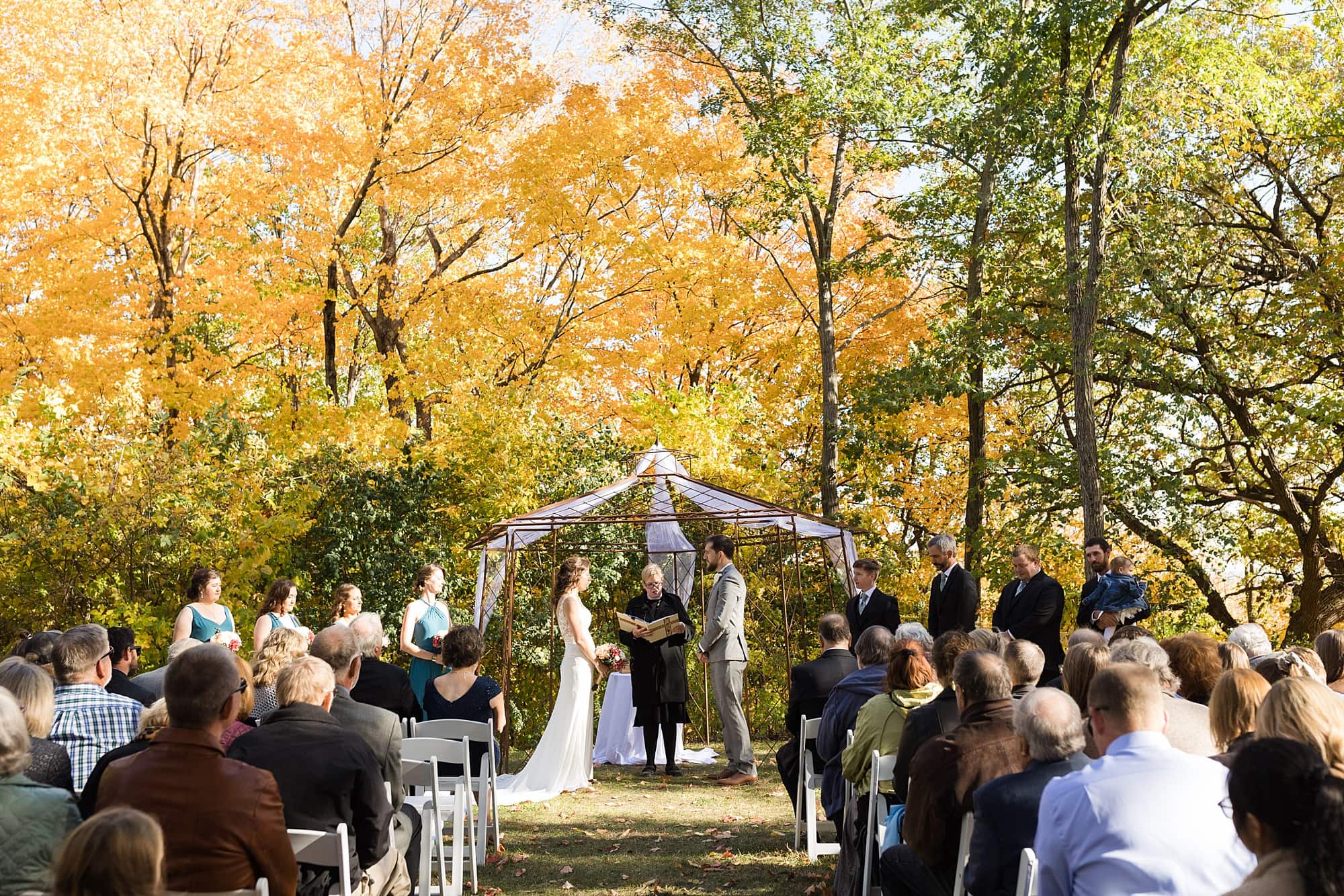 Bride and groom are surrounded by fall colors during their outdoor wedding ceremony at Bluff Creek Golf Course
