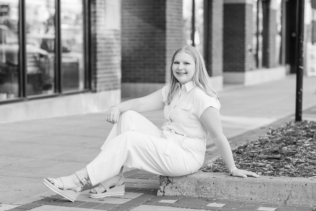 A girl in a cream colored jumpsuit sits on the sidewalk during her Downtown Fargo High School Senior session