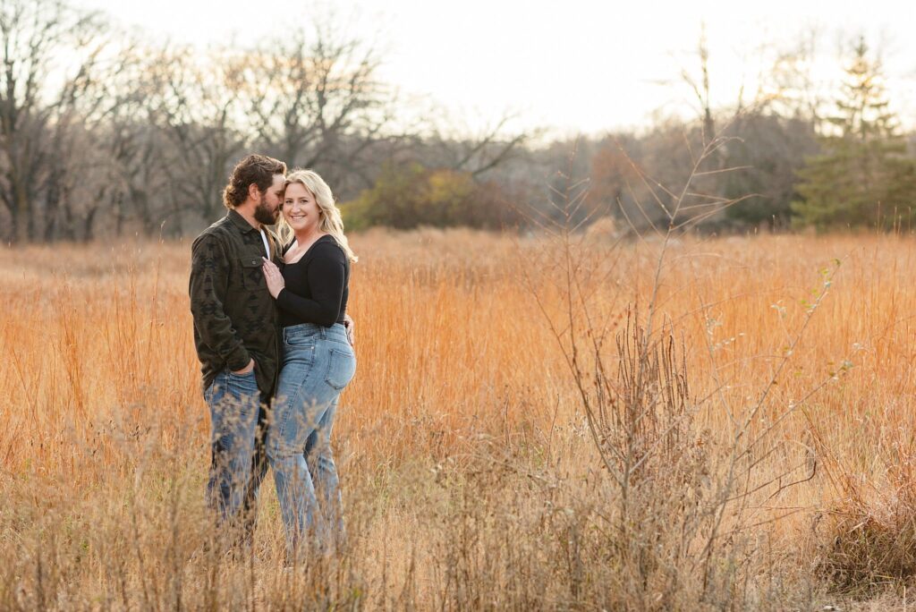 Engaged couple in jeans and dark tops smile in the orange tall grass field as the sun sets in Forest River Park